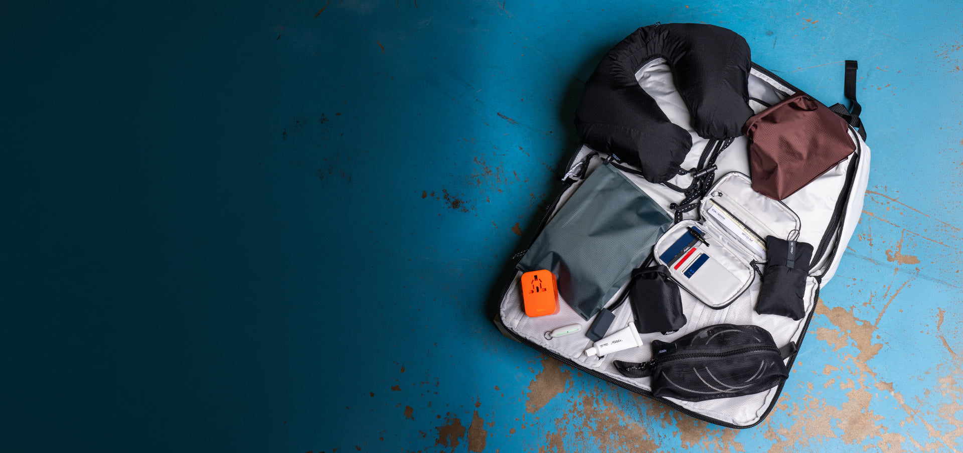 travel pack laying on blue floor with toiletry items, travel pillow, and bright orange power adapter