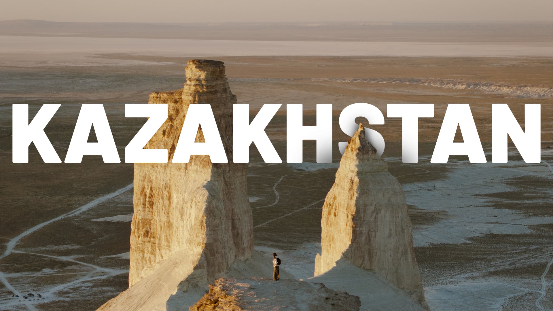 A travel-style scenic image of a vast desert landscape in Kazakhstan with two tall rock spires, a small person standing below, and large white text reading 'KAZAKHSTAN' across the center.