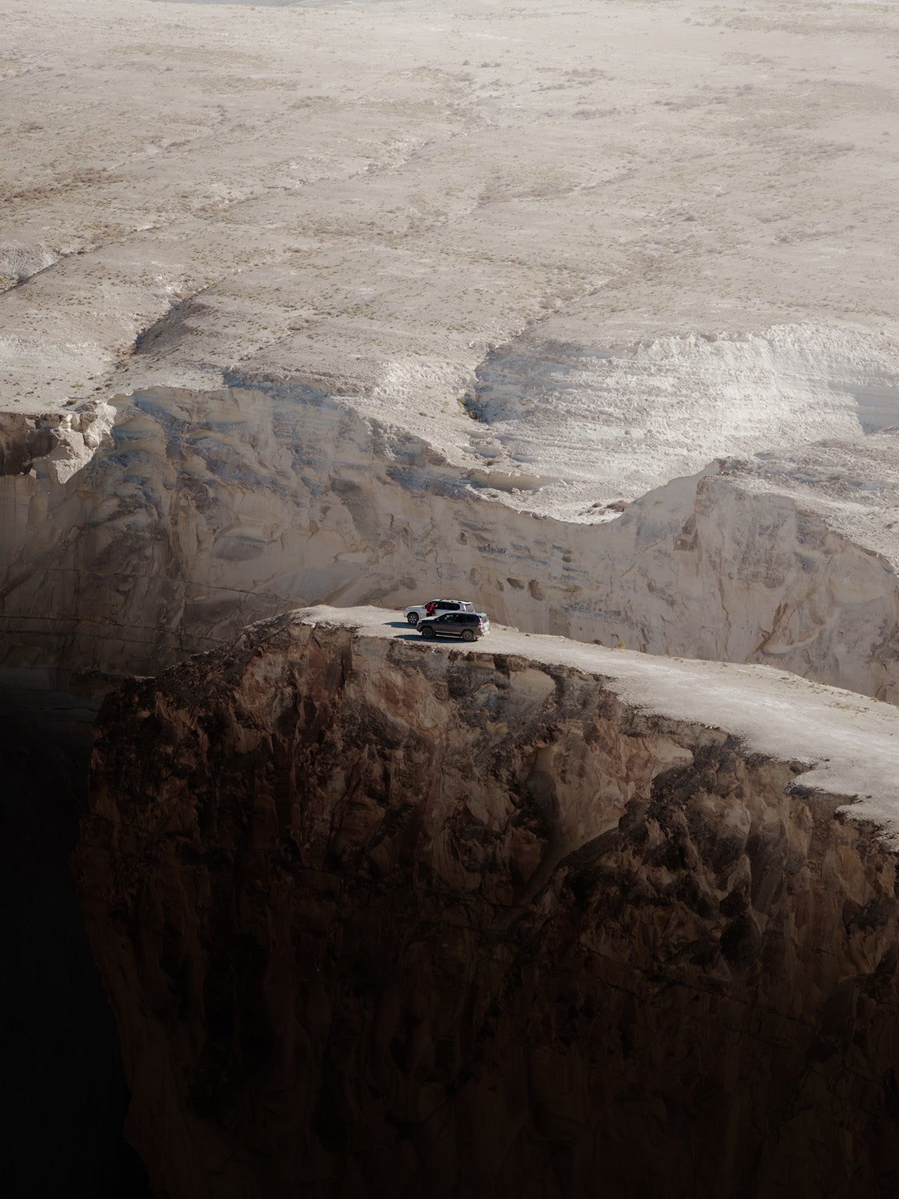 A small black vehicle parked on a narrow road during a Mangystau Road Trip along a high rocky cliff, overlooking a vast pale desert or glacier-like landscape.