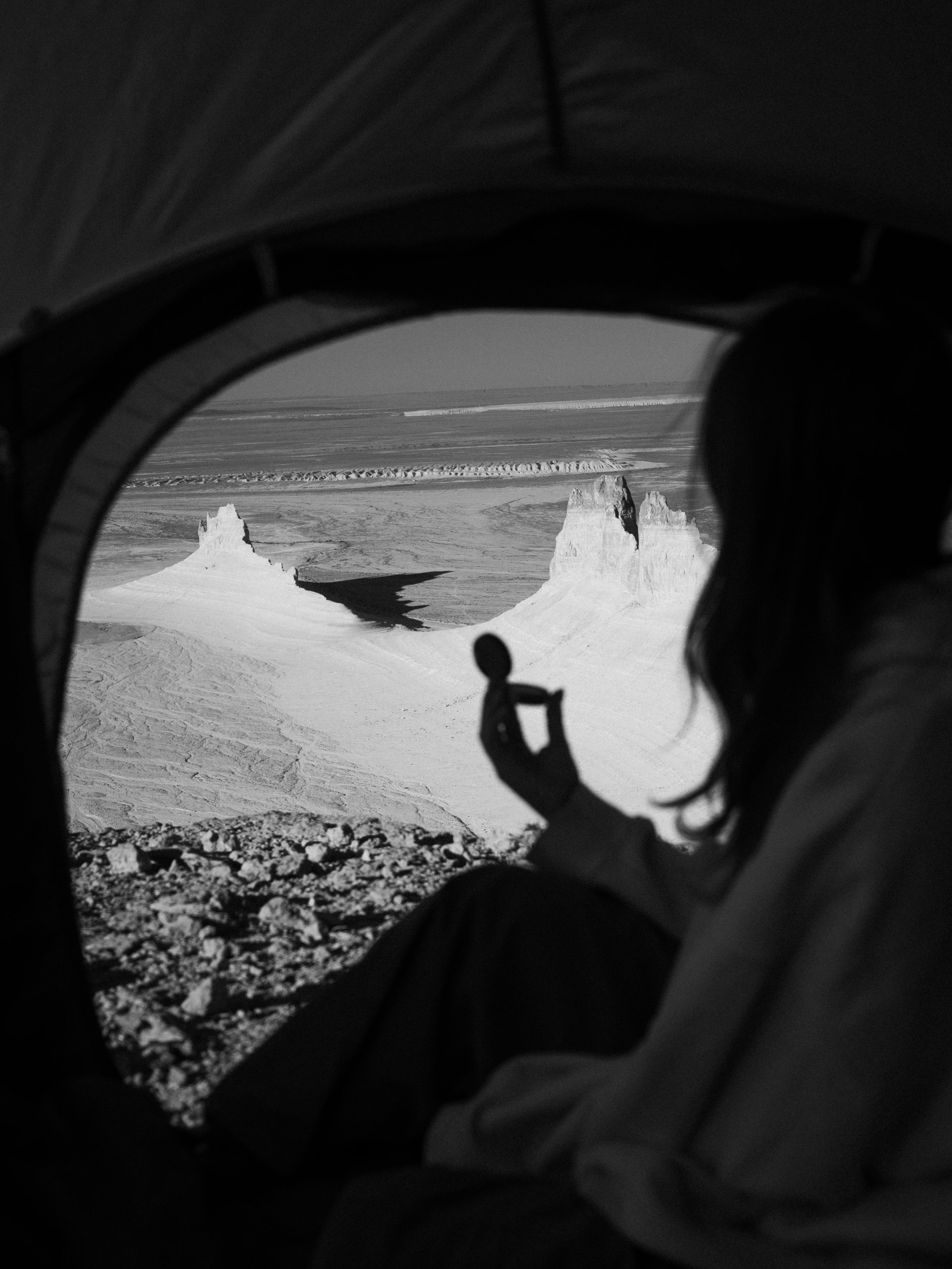 Black and white photo of a person sitting inside a tent, looking out at a dramatic rocky desert landscape with a small object in their hand.