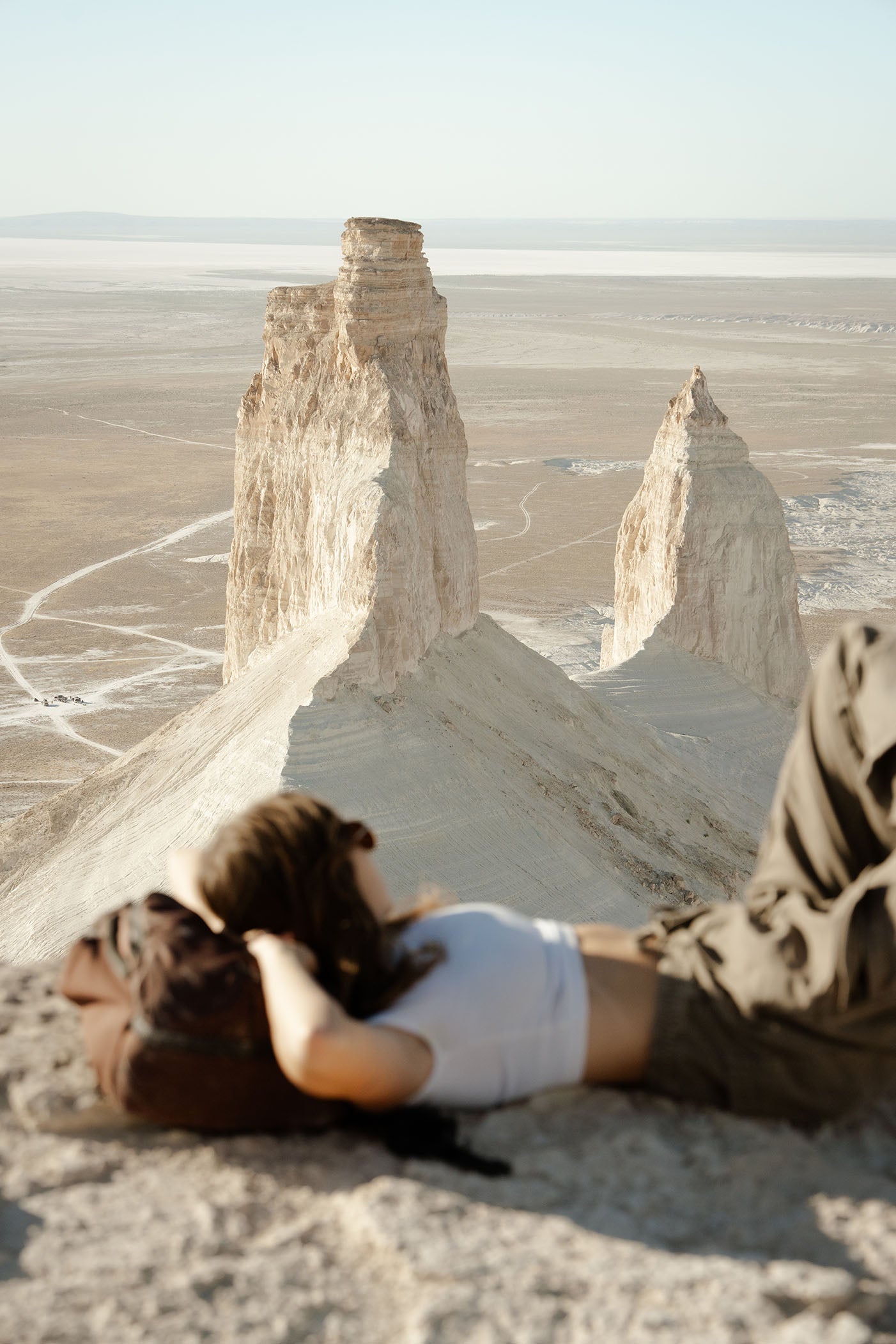 A woman lies on dark red travel backpack on a rocky desert ridge, looking toward large pale rock spires and a vast desert landscape under a clear sky.