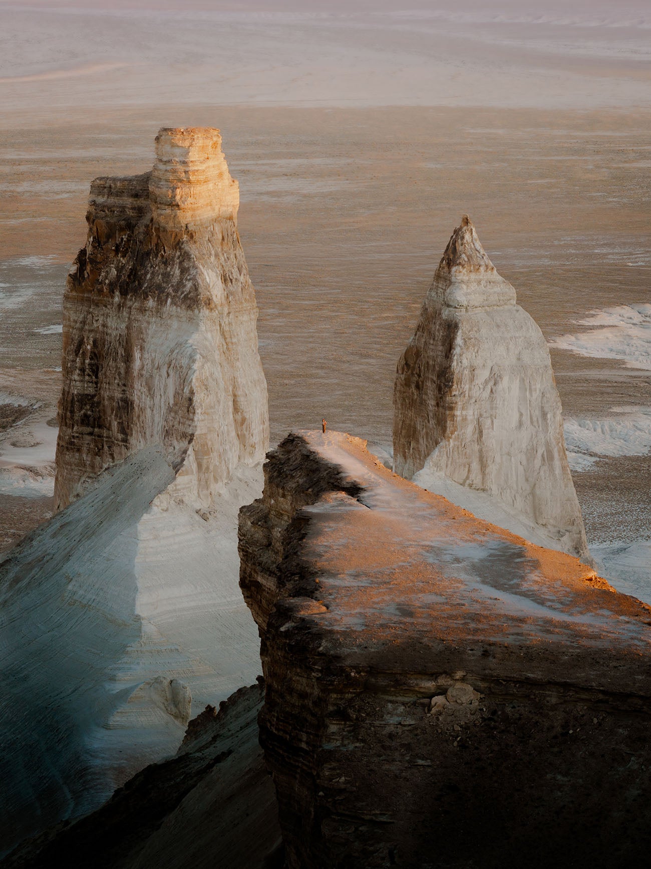 A dramatic landscape photograph of tall, slender rock spires called The Fangs of Bozzhyra rising from a flat, pale terrain under warm low light, with a dark rocky ledge in the foreground.