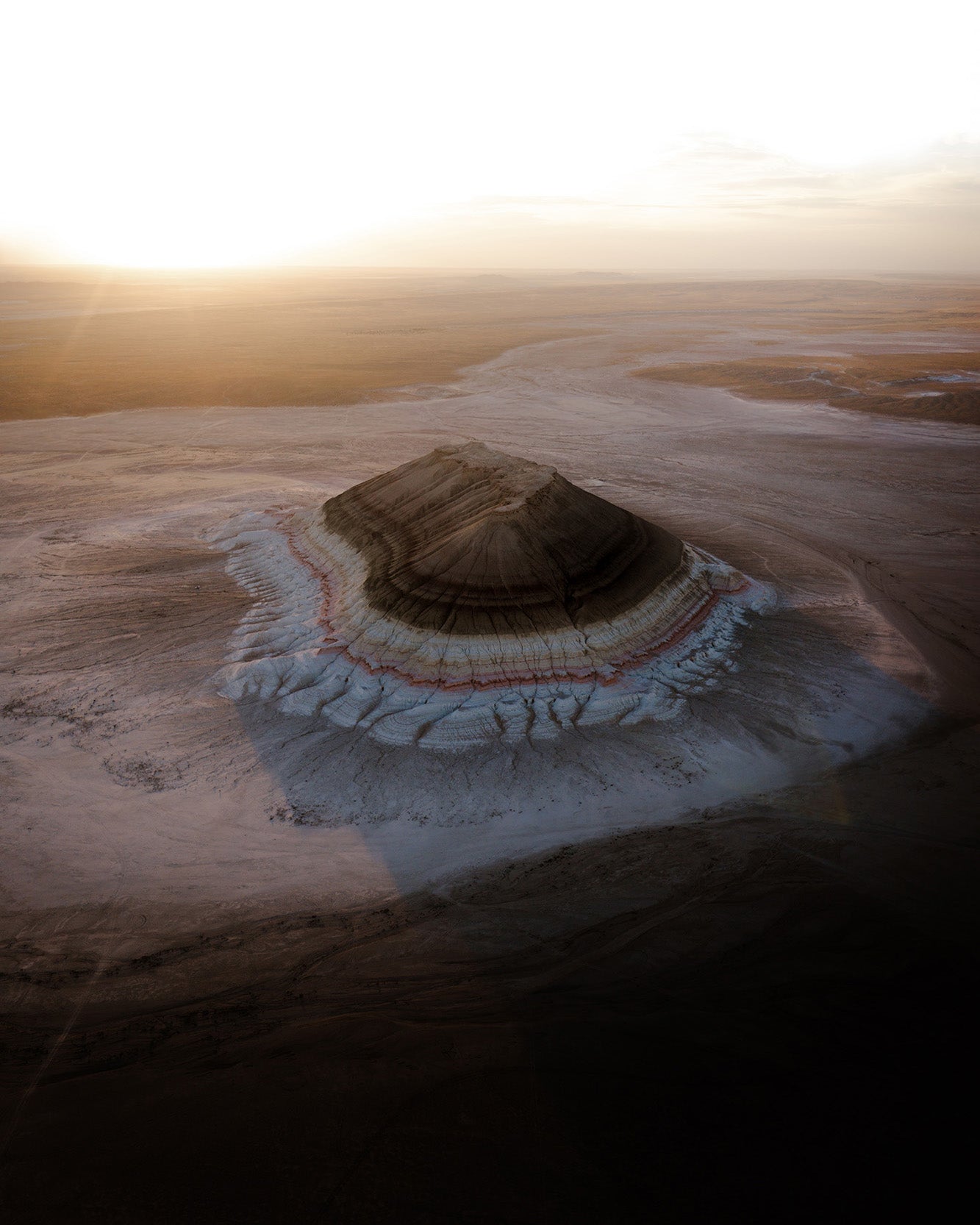 Aerial view of a layered volcanic or mineral formation surrounded by pale salt flats or desert terrain under a bright hazy sky.
