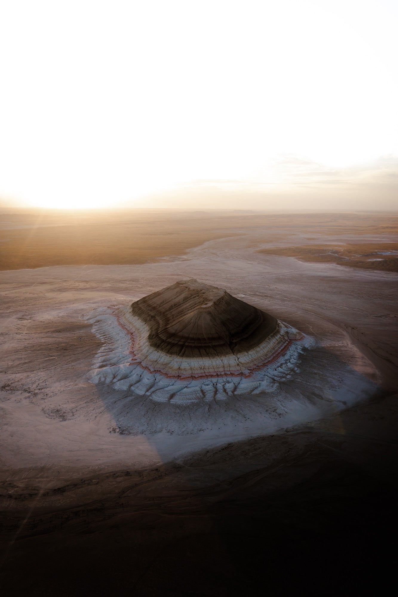 Aerial view of a circular salt flat called tiramisu canyon in Kazakhstan with concentric ring patterns in white, tan, and dark brown, surrounded by a bright sunlit horizon.