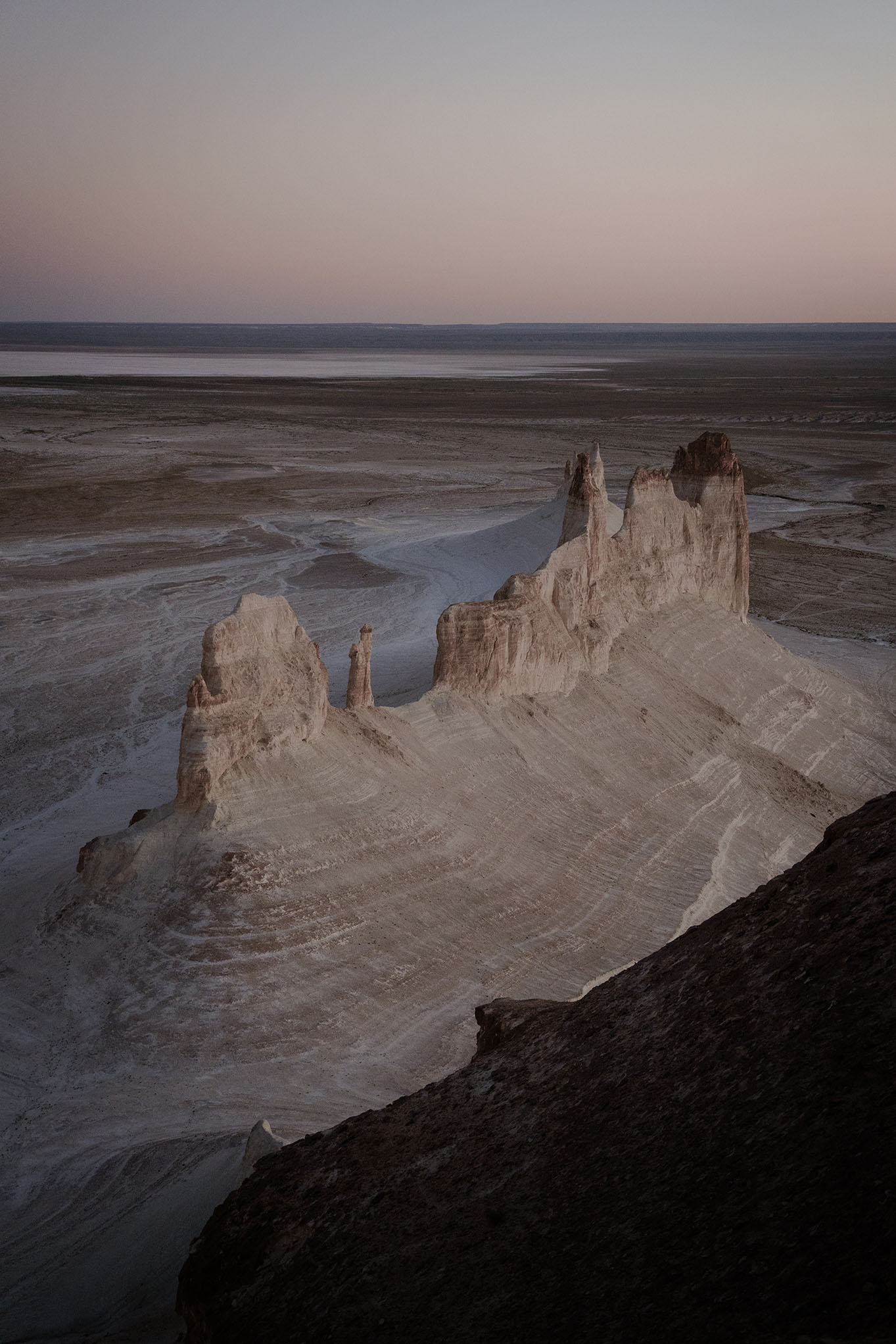 A dramatic landscape photograph of white rock spires and salt flats under a pale twilight sky, with a dark foreground slope.