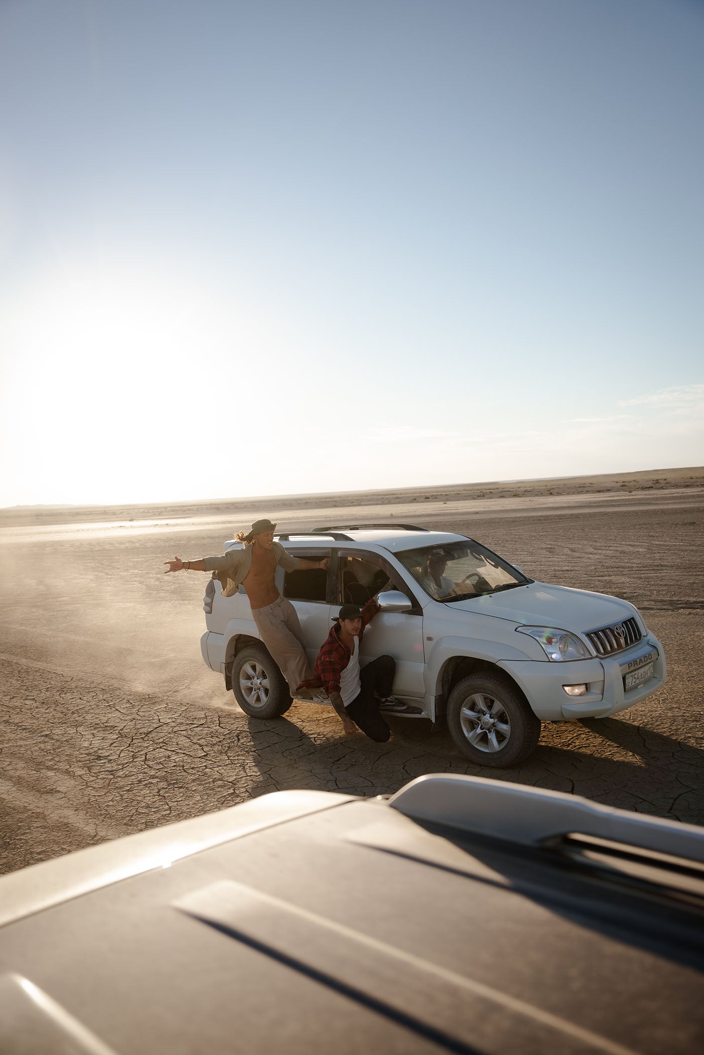 A white SUV parked on a dusty desert road with two people beside it, one leaning out with arms wide and the other sitting in the open door. The photo is taken from a vehicle in the foreground under bright sunlight.
