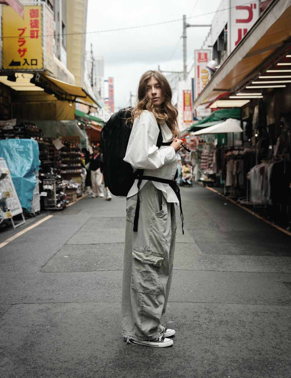 A woman stands on a narrow urban Japanese street lined with market stalls and storefronts, wearing a white long-sleeve top, loose light gray cargo pants, a black backpack, and black low-top sneakers.