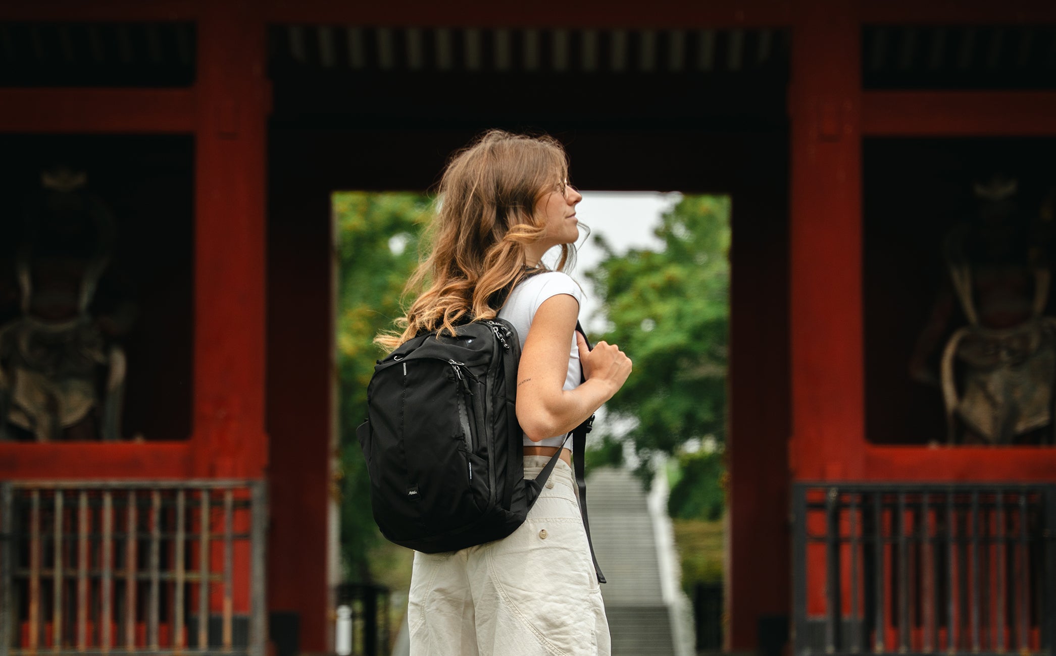 A woman stands in profile wearing white clothes and a small black backpack, positioned in front of a red traditional Japanese entrance with green foliage visible in the background.