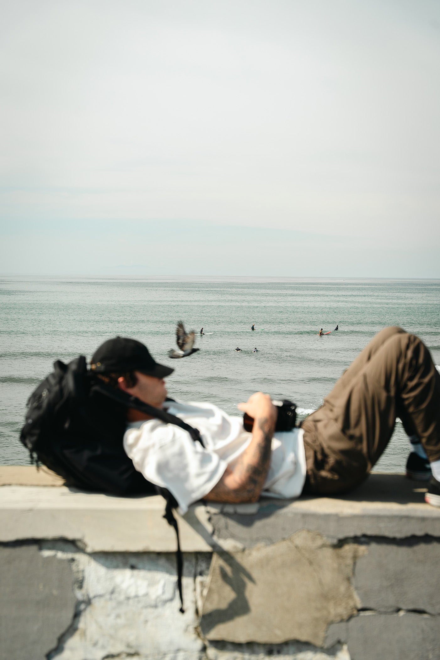 A man resting on a black backpack, wearing a white t-shirt, brown pants, and a black cap lies on a stone wall by the ocean, holding a camera. A black backpack rests beside him, with birds and surfers visible in the water under a pale sky.