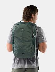 A man viewed from behind wearing a dark green backpack on his back, paired with a gray t-shirt, against a light gray studio background.