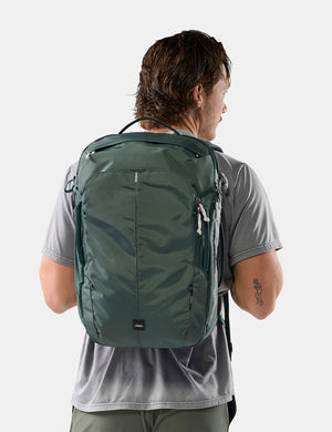 A man viewed from behind wearing a dark green backpack on his back, paired with a gray t-shirt, against a light gray studio background.