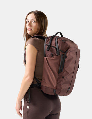 A woman wearing a brown sleeveless top and matching leggings is shown from the back wearing a large garnet backpack with black straps and zipper details against a light gray studio background.