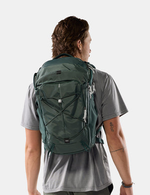 A man wearing a dark green technical backpack, viewed from behind to show the fit and design, against a light gray studio background.