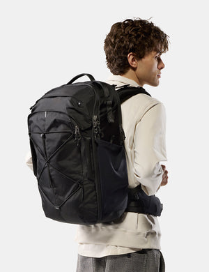 A man wearing a cream-colored hoodie and gray pants is shown from the back and side, carrying a large black technical backpack against a light gray studio background.