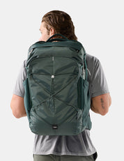 A man viewed from behind wearing a dark green technical backpack with multiple zippered compartments and compression straps, set against a plain light gray studio background.