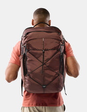 A man viewed from behind wearing a large garnet technical backpack with black compression straps and zippers, over a coral t-shirt, against a light gray background.