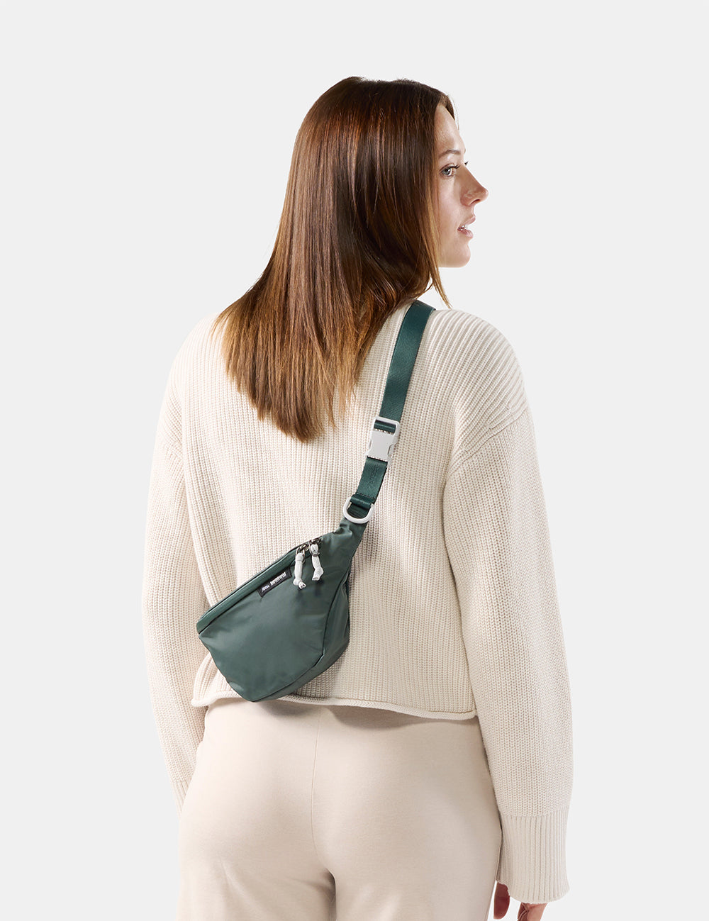 A woman wearing a cream ribbed sweater and beige pants, shown from the back and side profile, wearing a dark green nylon fanny pack 