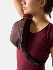A close-up of a model wearing a dark maroon and black sling bag across their chest, paired with a burgundy t-shirt against a light gray background.