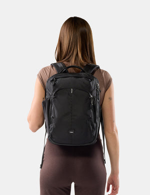 A woman viewed from behind wearing a black backpack against a light gray studio background.