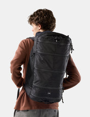 A man wearing a large black backpack, photographed from behind against a light gray studio background.