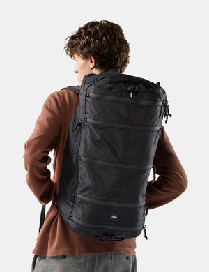 A man wearing a large black backpack, photographed from behind against a light gray studio background.