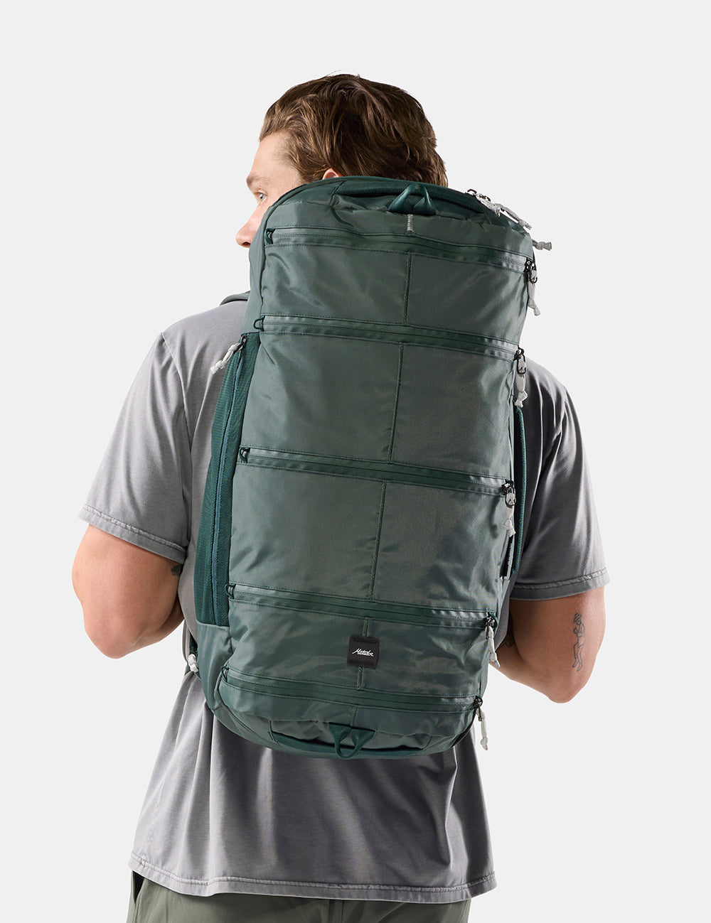 A man wearing a gray t-shirt is shown from behind, carrying a large, dark green technical backpack against a light gray studio background.