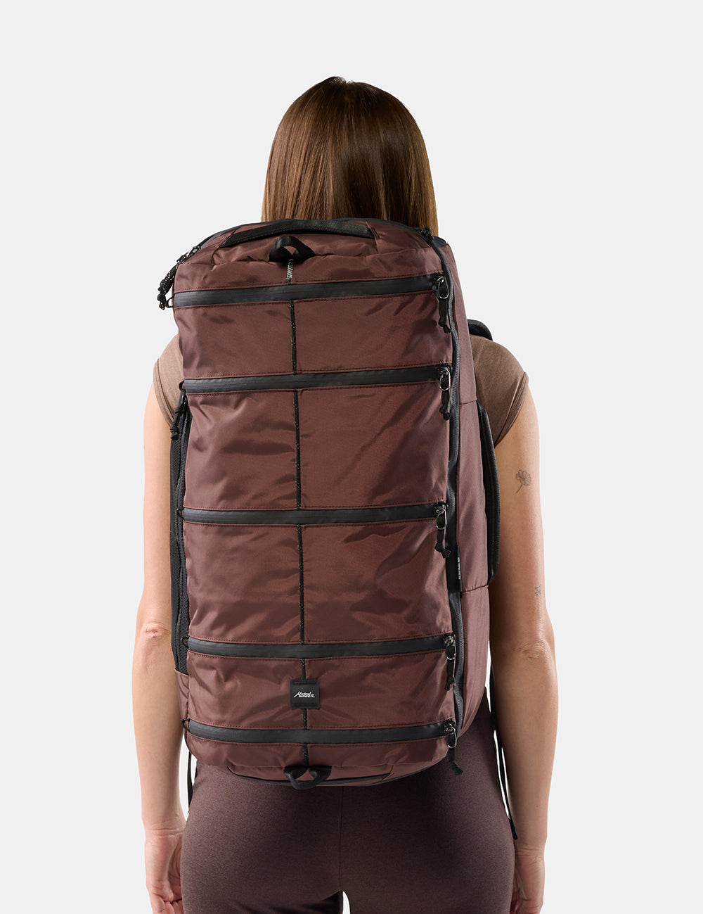 A woman viewed from behind wearing a large garnet segmented backpack against a light gray studio background.
