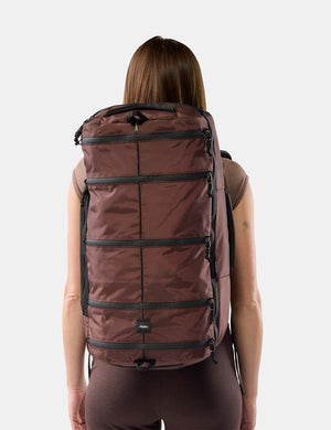 A woman viewed from behind wearing a large garnet segmented backpack against a light gray studio background.