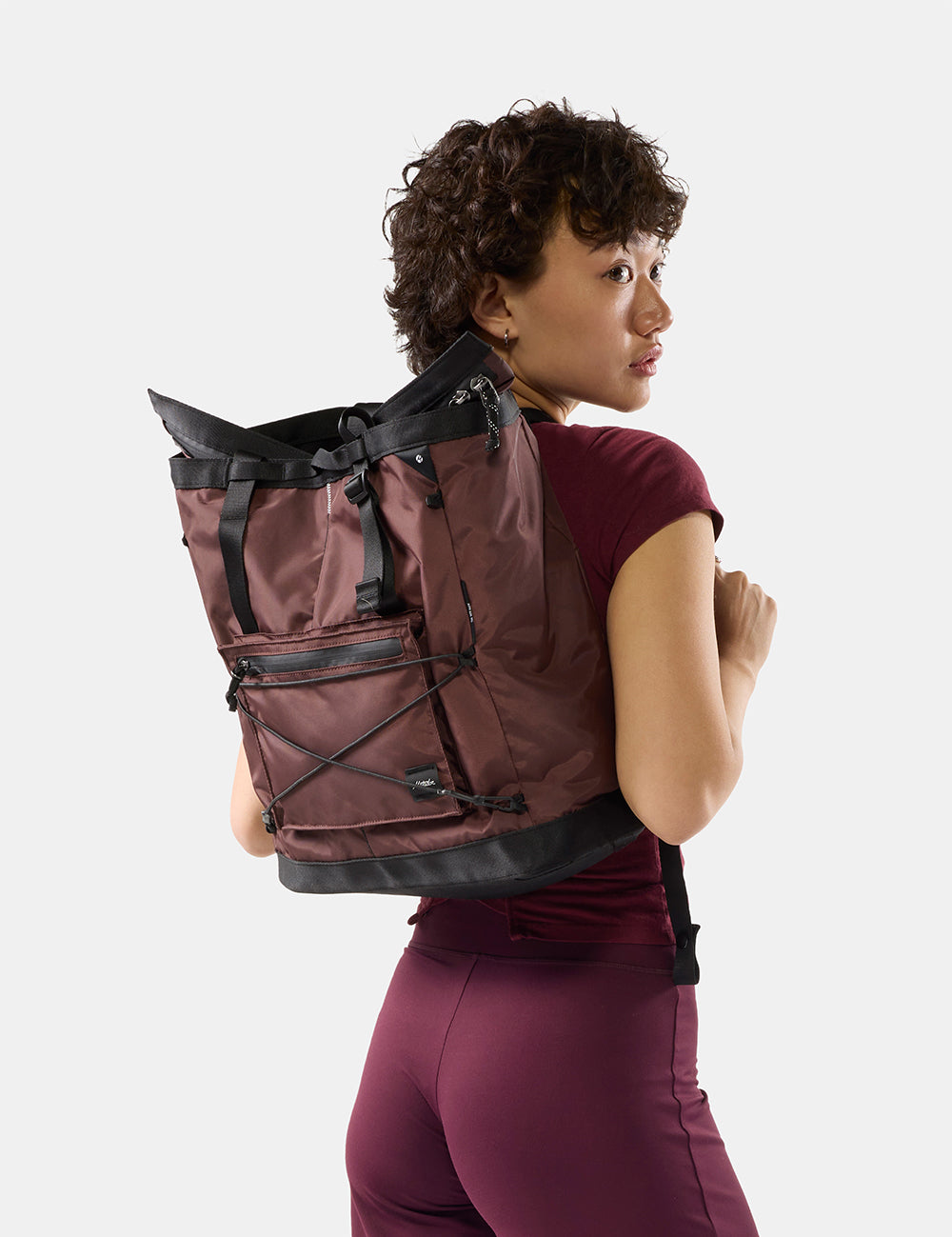 A woman wearing a garnet backpack with black trim and straps, shown from a side-back angle against a light gray studio background.