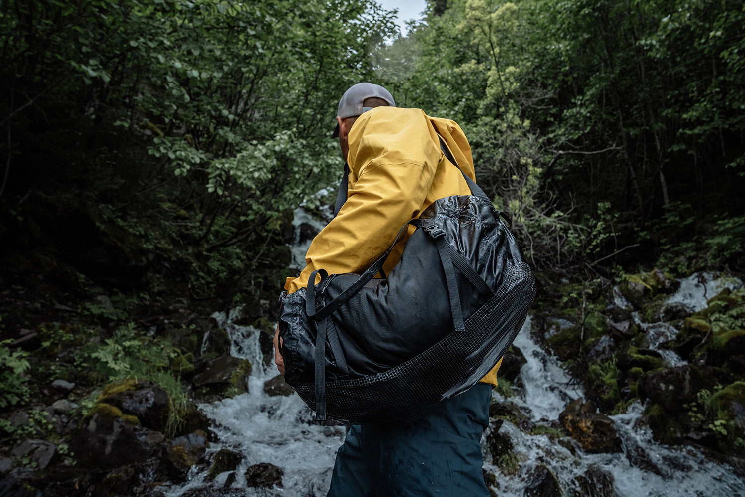 Person in a yellow raincoat carrying a black waterproof packable duffle bag through a forest with a stream.