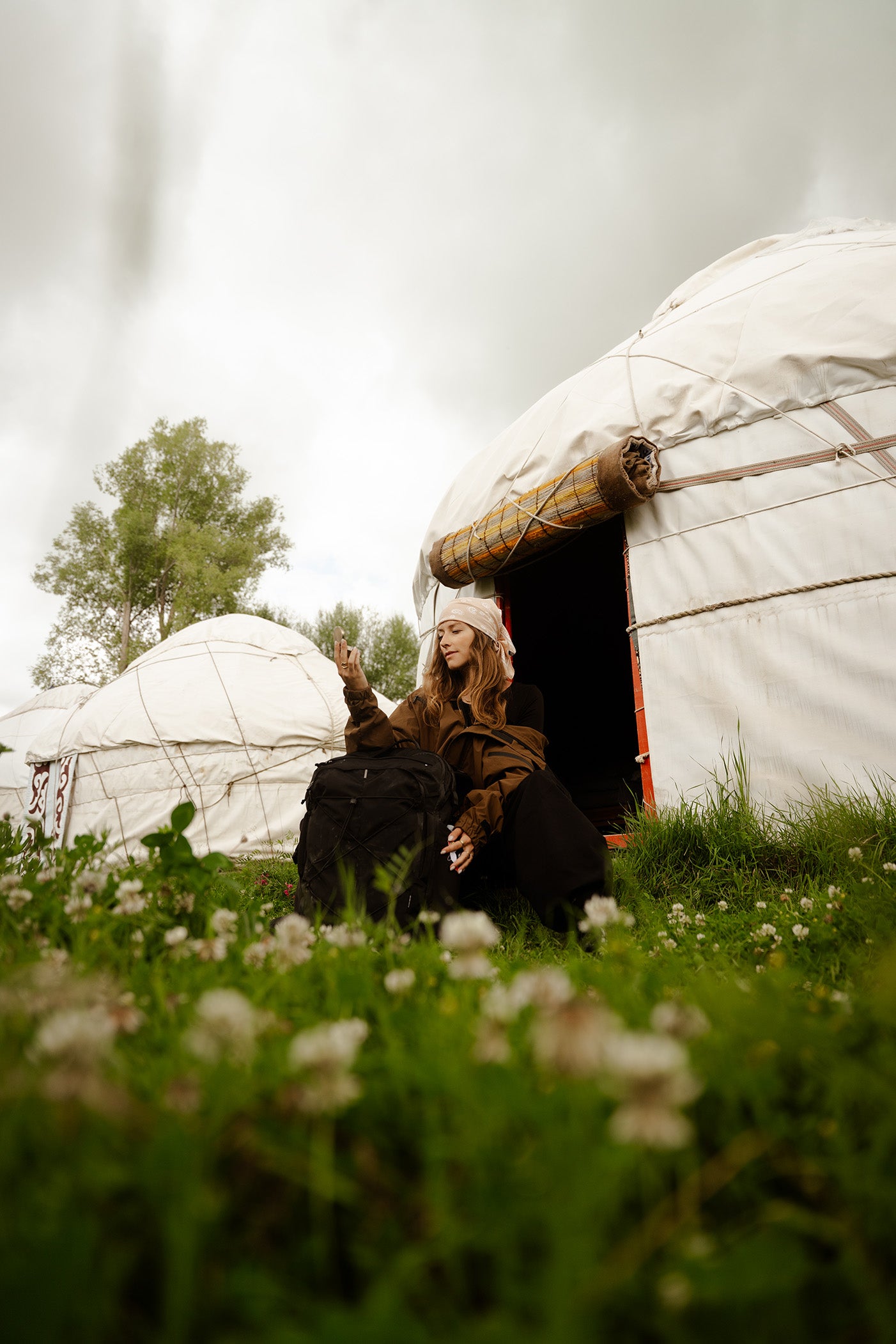 A woman in a brown coat and white hat sits on the grassy entrance of a white yurt in an outdoor camp with a black travel bag.