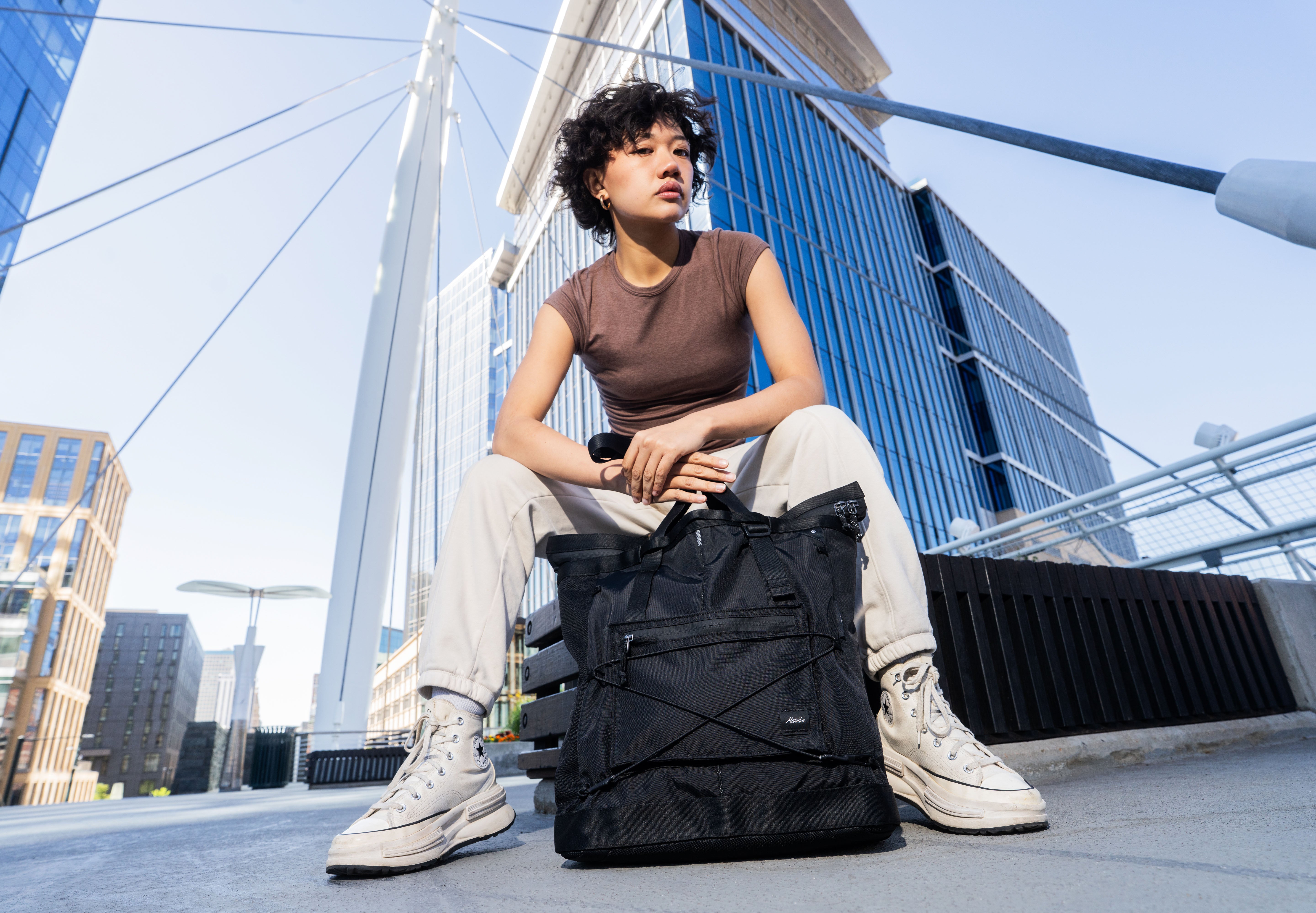person sitting on a bench with a black tote bag and a city scape behind her with tall buildings and a bridge