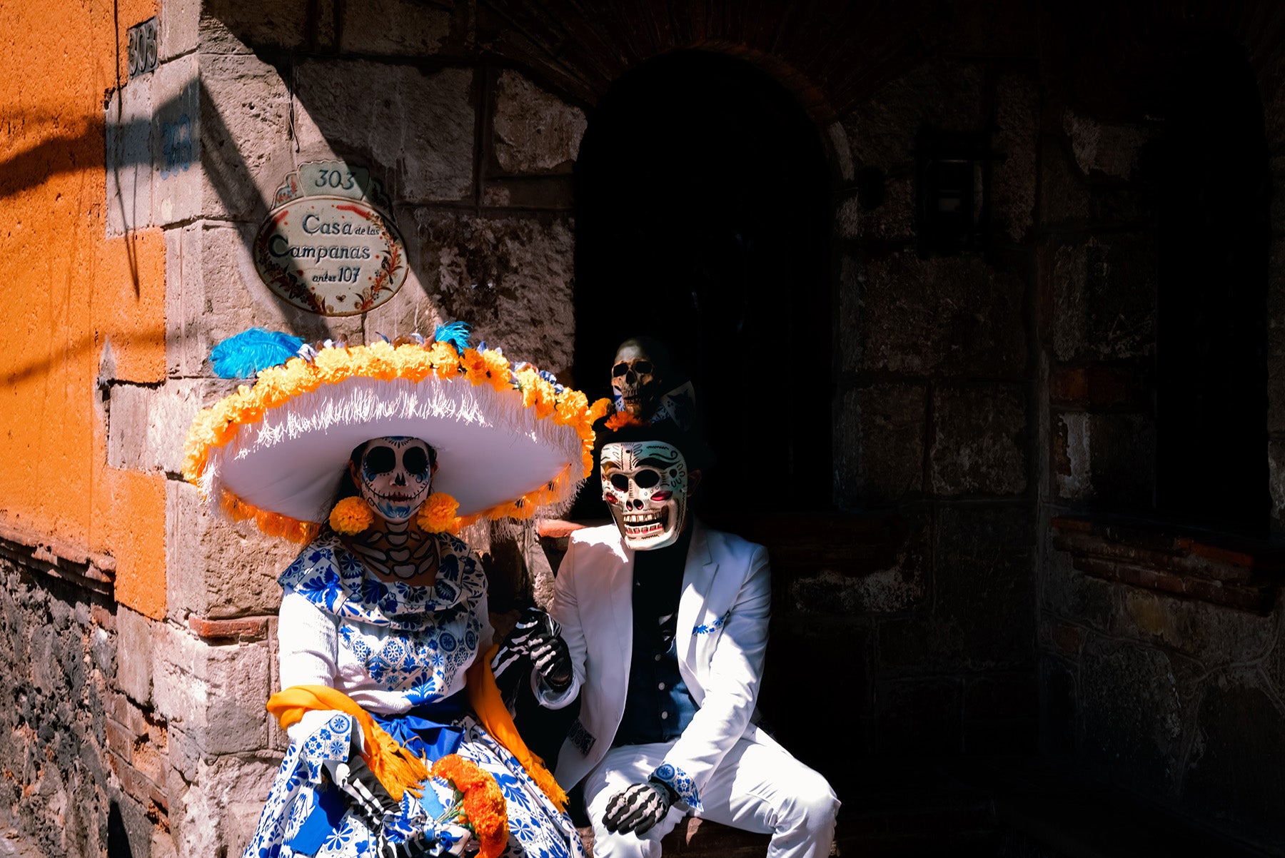 Two people dressed in Day of the Dead (Día de los Muertos) costumes posing in front of a rustic stone wall and arched doorway. The person on the left wears a large white sombrero with orange marigolds, a blue and white patterned dress, and orange flower trim.