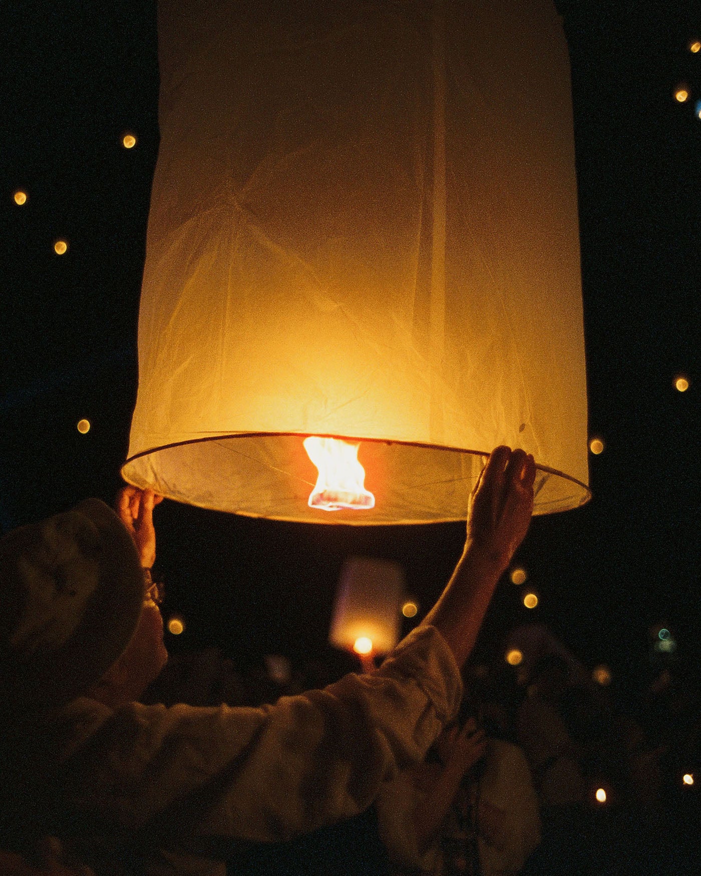 A person at night holds up a large sky lantern with a small flame inside, with other lanterns glowing in the dark background.