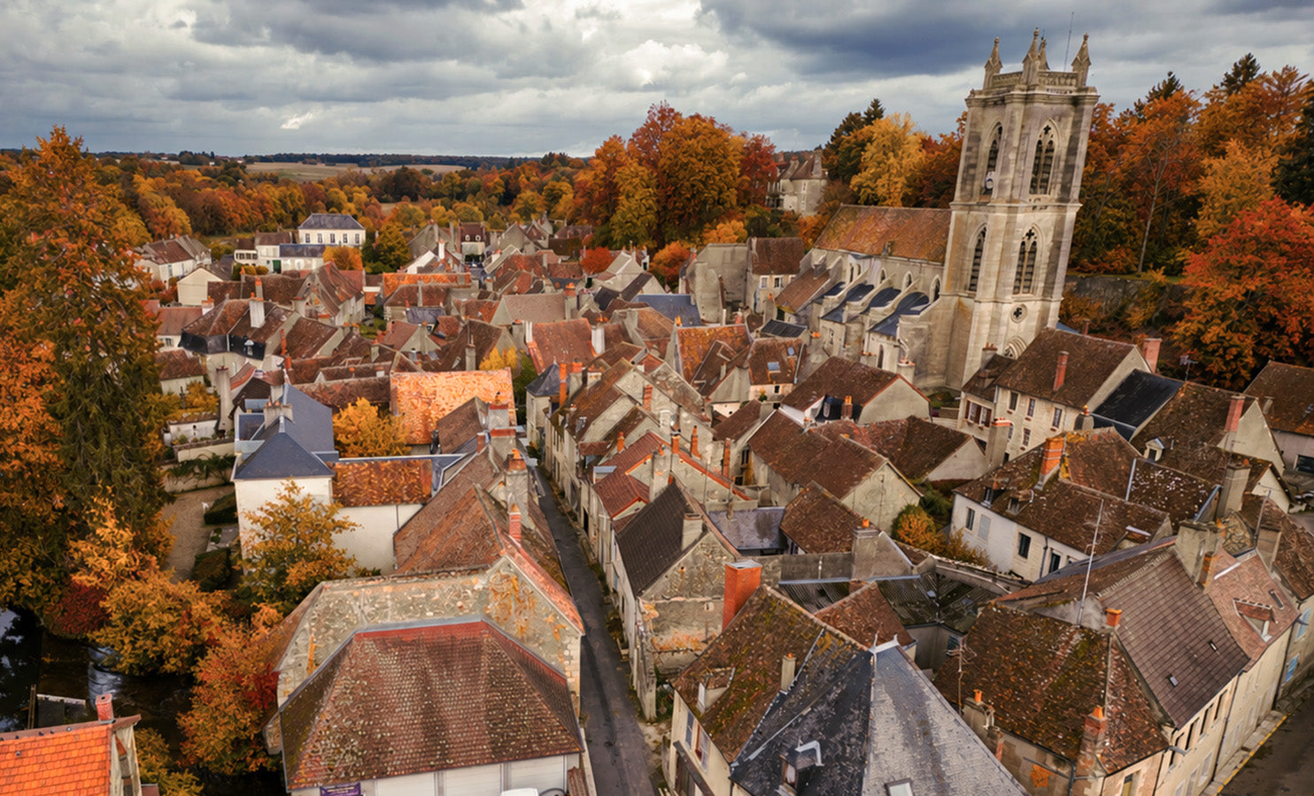 Aerial view of a historic European town with dense stone buildings, terracotta and gray rooftops, a tall church tower, and autumn trees in orange, yellow, and green under a cloudy sky.