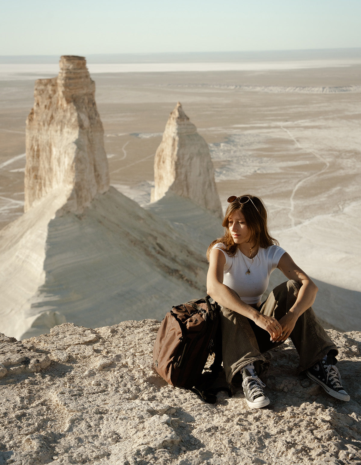 A woman sits on a rocky desert cliffside next to a dark red travel backpack with large pale rock spires in the background. She is wearing a white sleeveless top, loose brown pants, and dark sneakers.