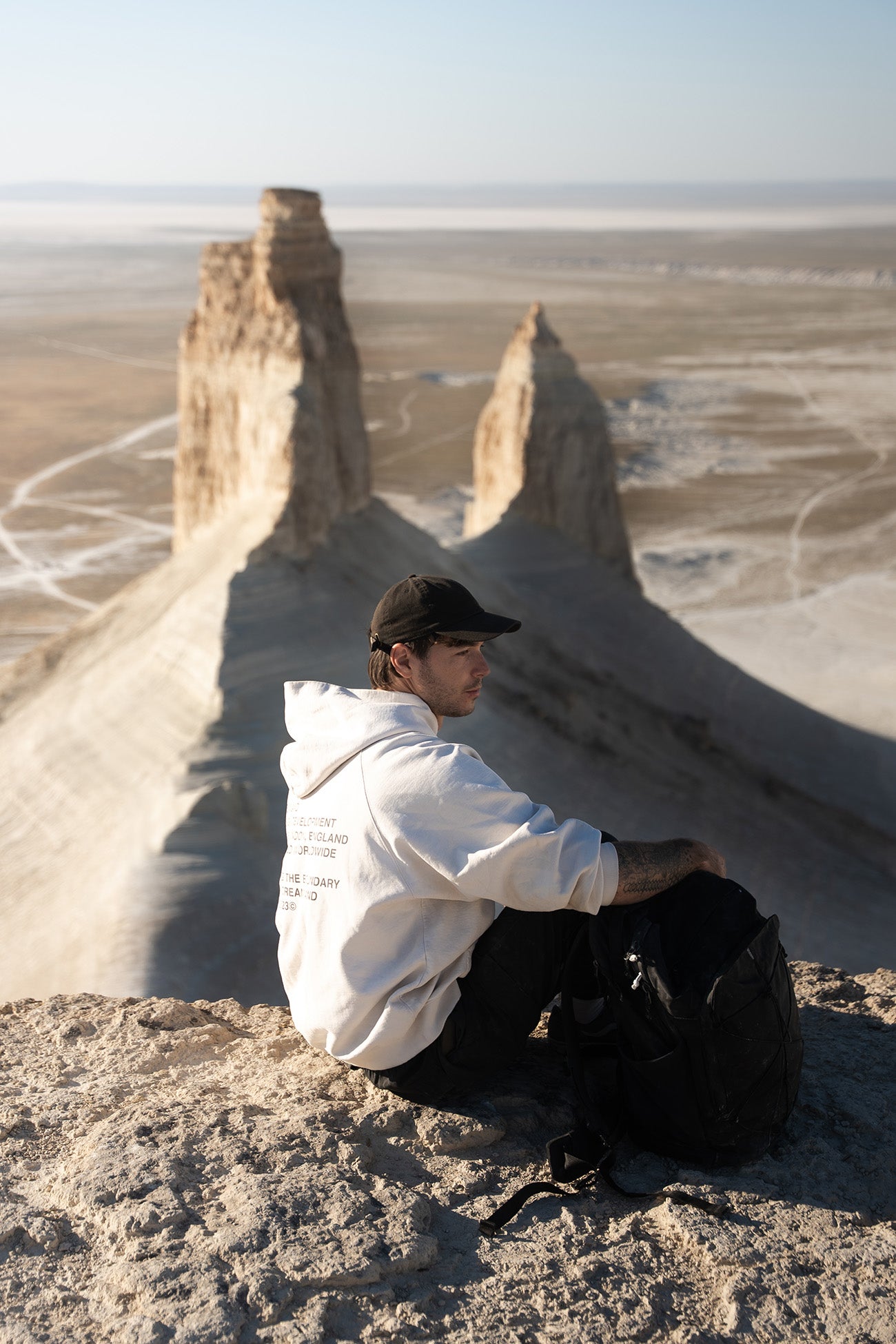 A man wearing a white hoodie, black pants, and a black cap sits on a rocky desert cliffside with dramatic sandstone spires in the background.