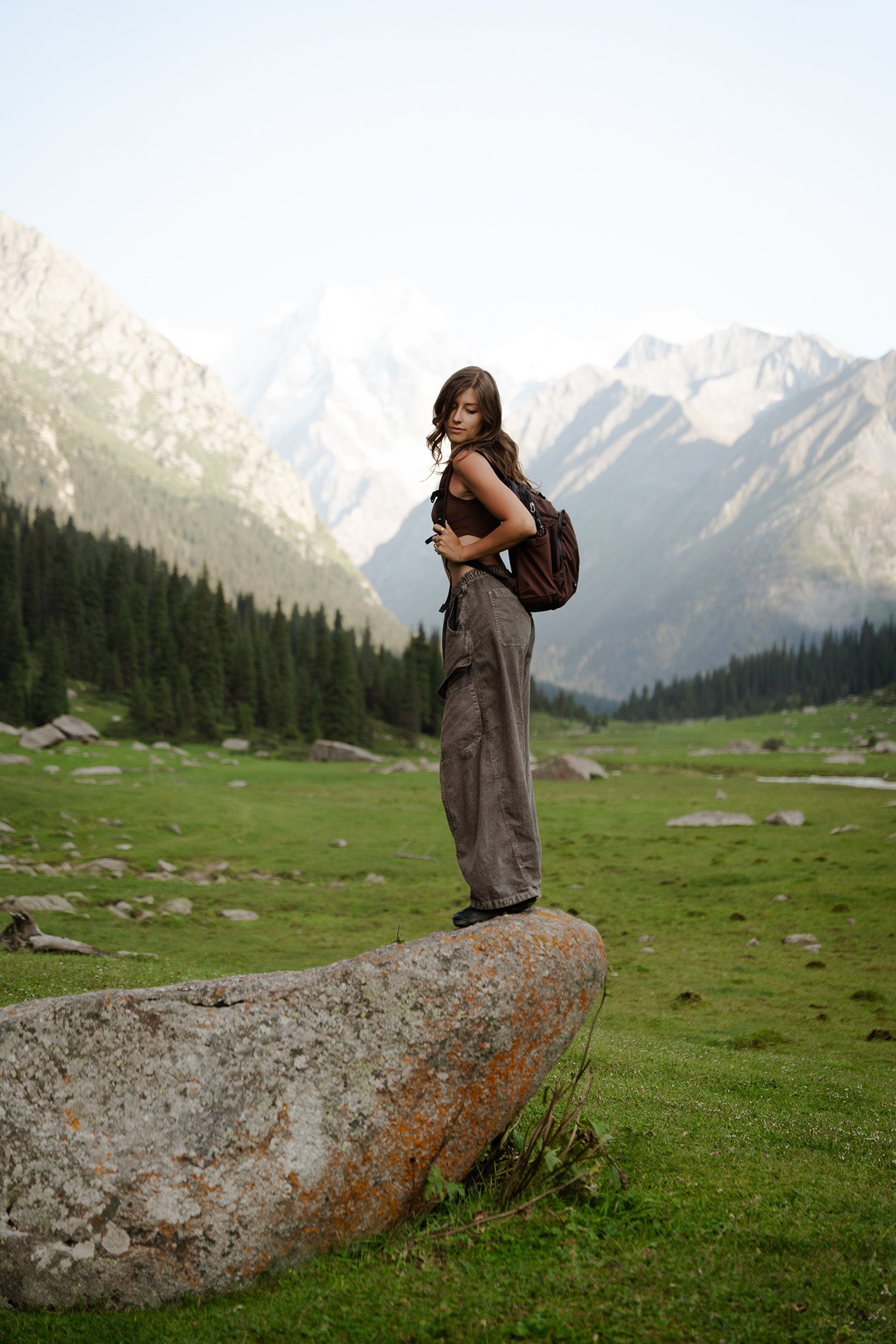 A woman stands on a large rock in a mountain meadow, wearing a dark brown sleeveless top, loose brown pants, and carrying a garnet 18L travel backpack, with snow-capped mountains and pine trees in the background.