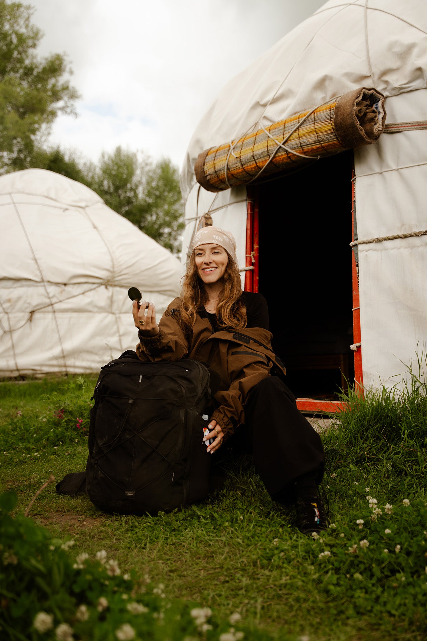 A woman sitting on the grass in front of a white yurt, holding a compact mirror by a black backpack and wearing a brown jacket, black pants, and a cream beanie, with another yurt and trees in the background.