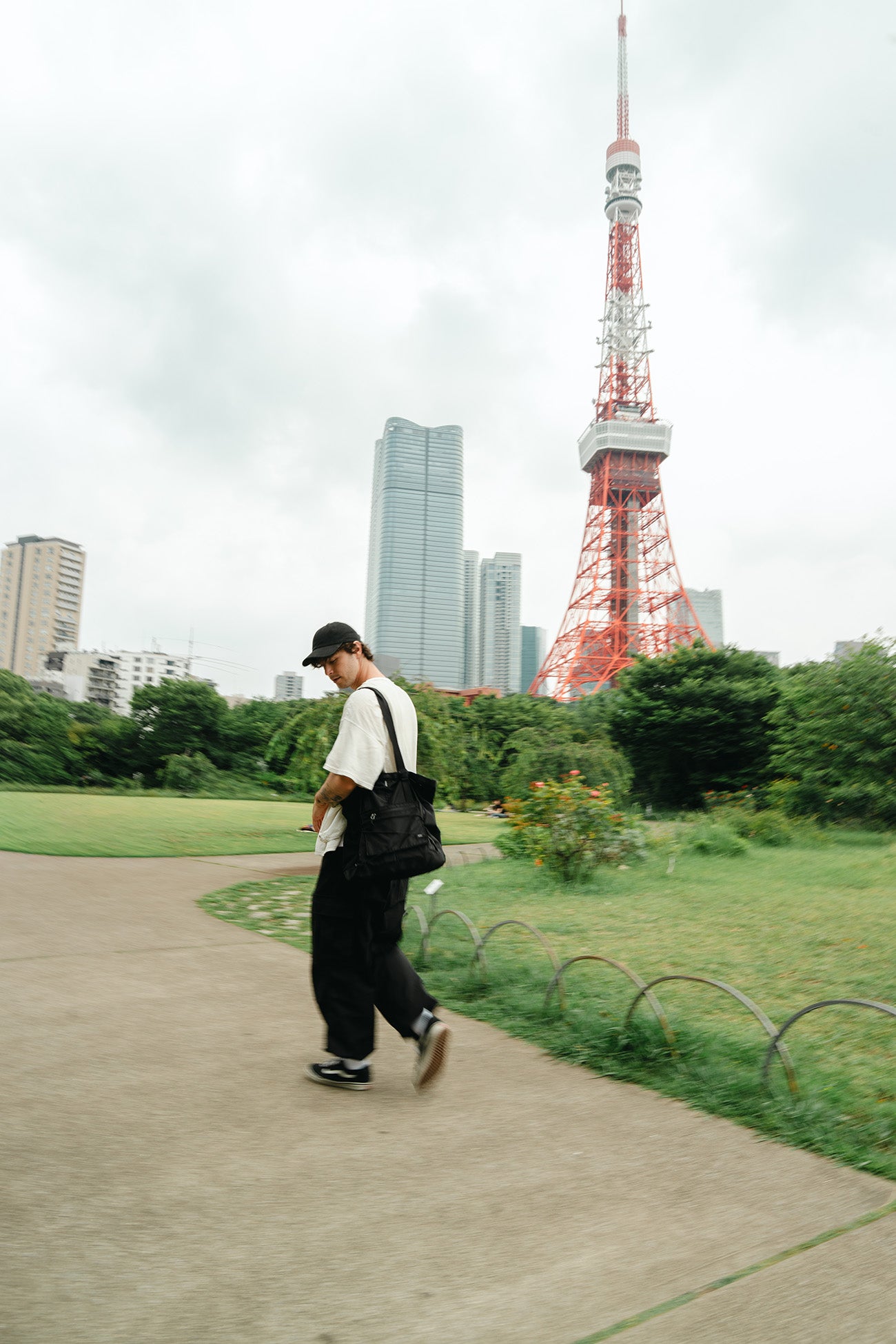 A man walking on a park path carrying a black tote bag over his shoulder, with the Tokyo Tower and city skyline in the background under an overcast sky.