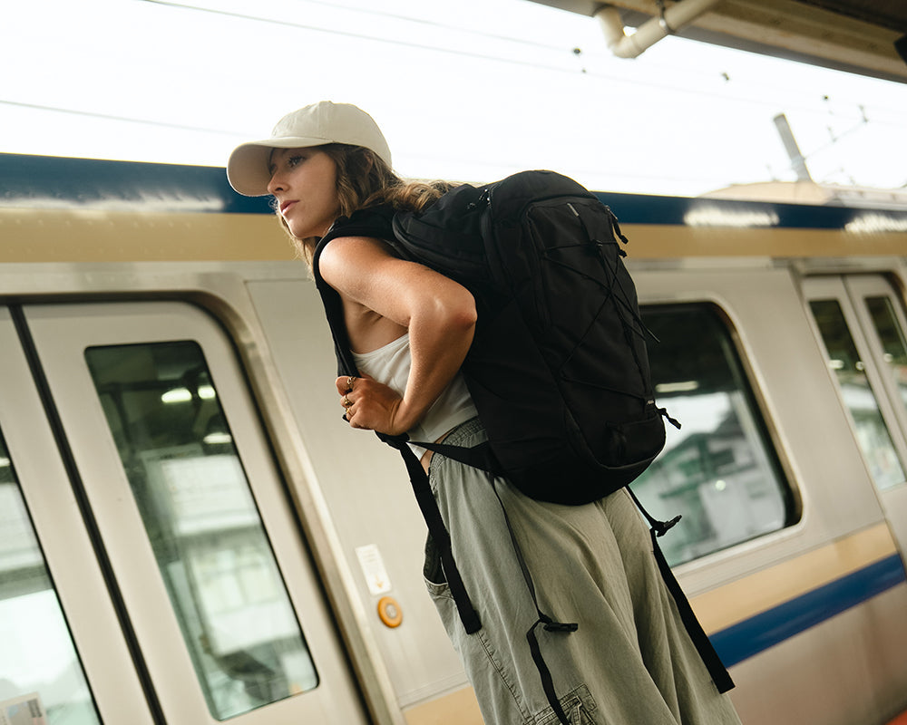 A woman stands on a train platform next to a silver commuter train, wearing a beige cap, a white tank top, light gray wide-leg cargo pants, and a large black backpack.
