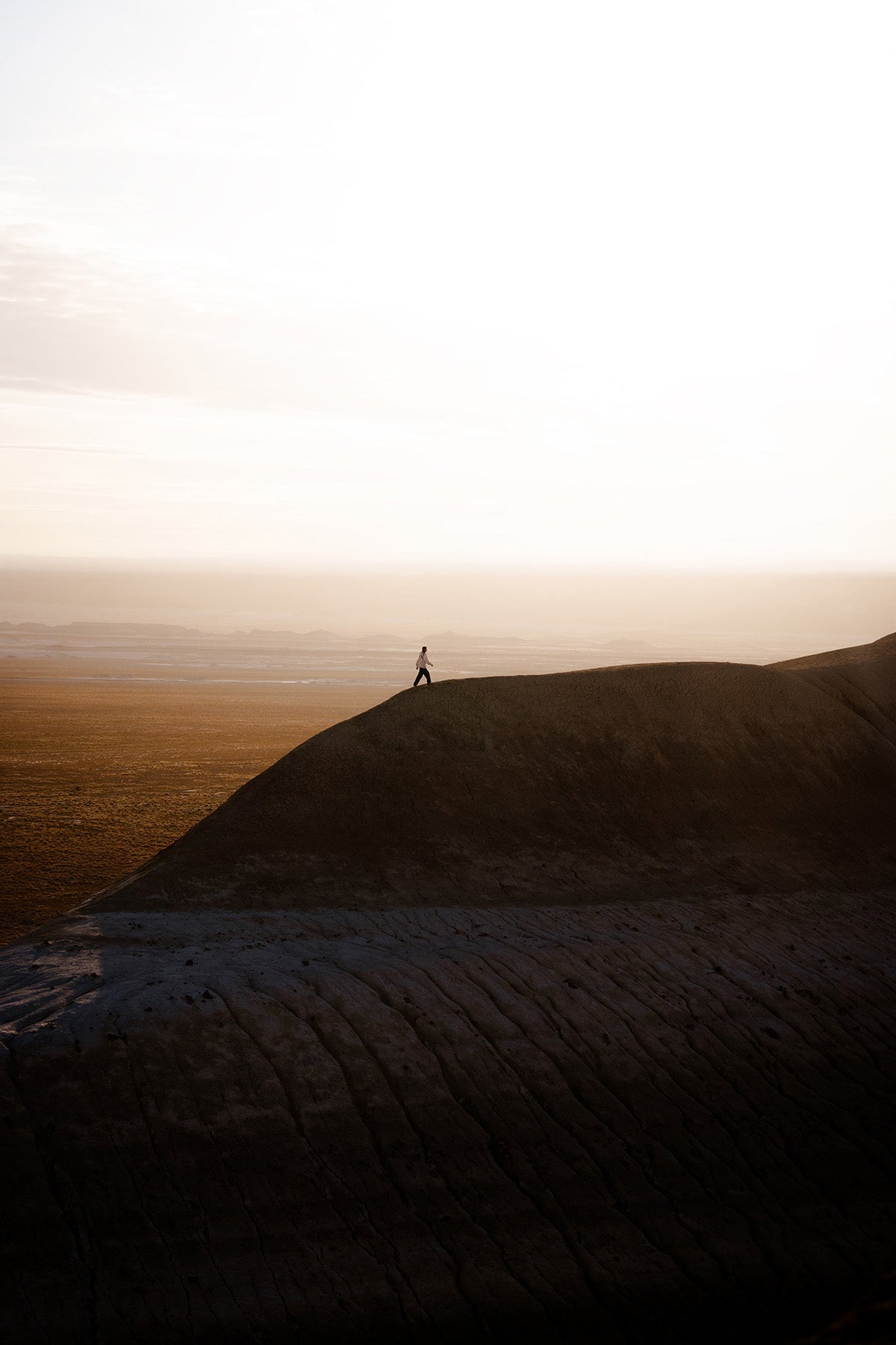 A silhouetted person stands at the crest of a large dark hill in a vast desert landscape during golden hour, with a bright hazy sky filling the upper half of the frame.
