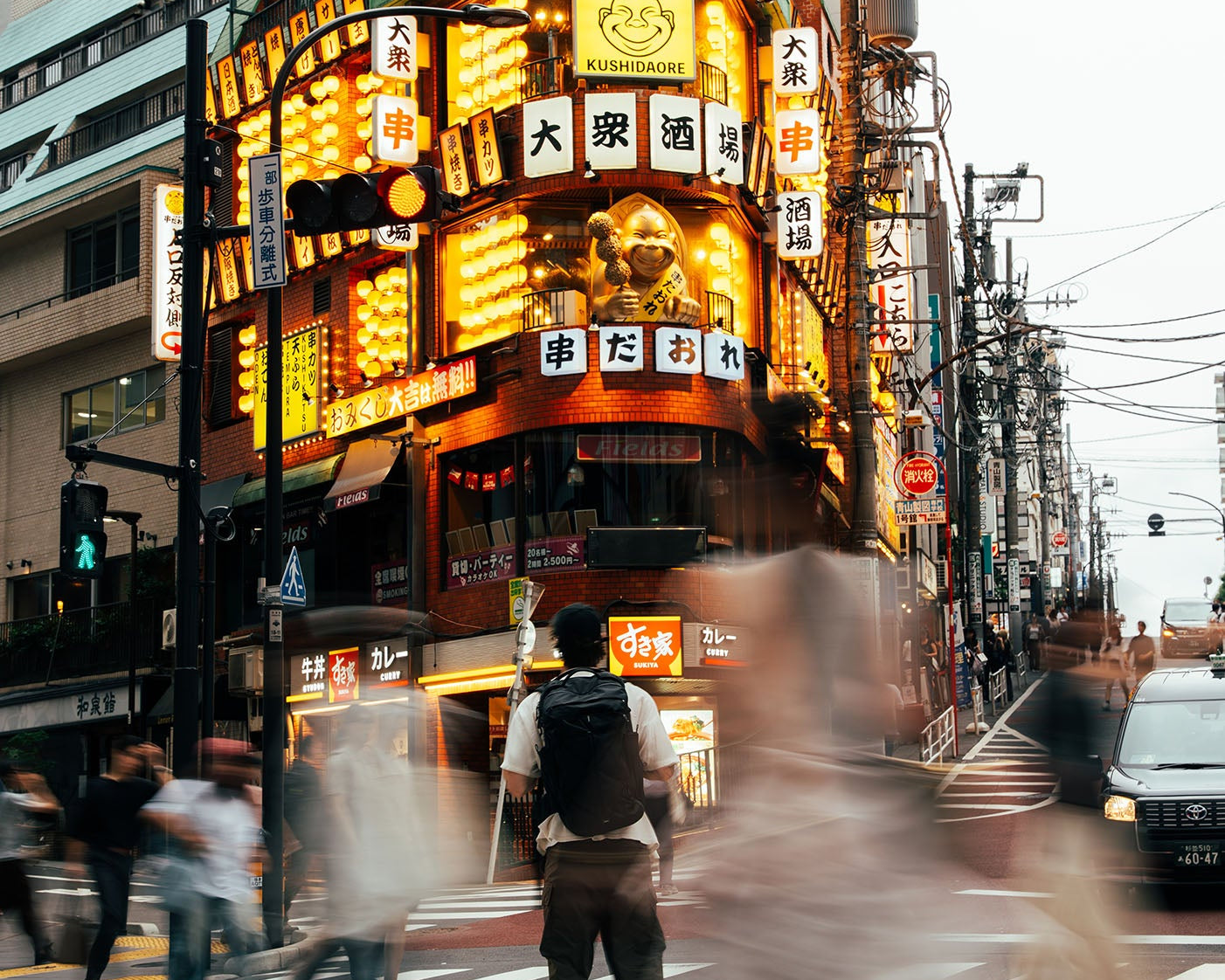 A bustling Japanese city street at dusk with a brightly lit ornate building, neon signs, pedestrians crossing a crosswalk, and motion blur in the foreground.