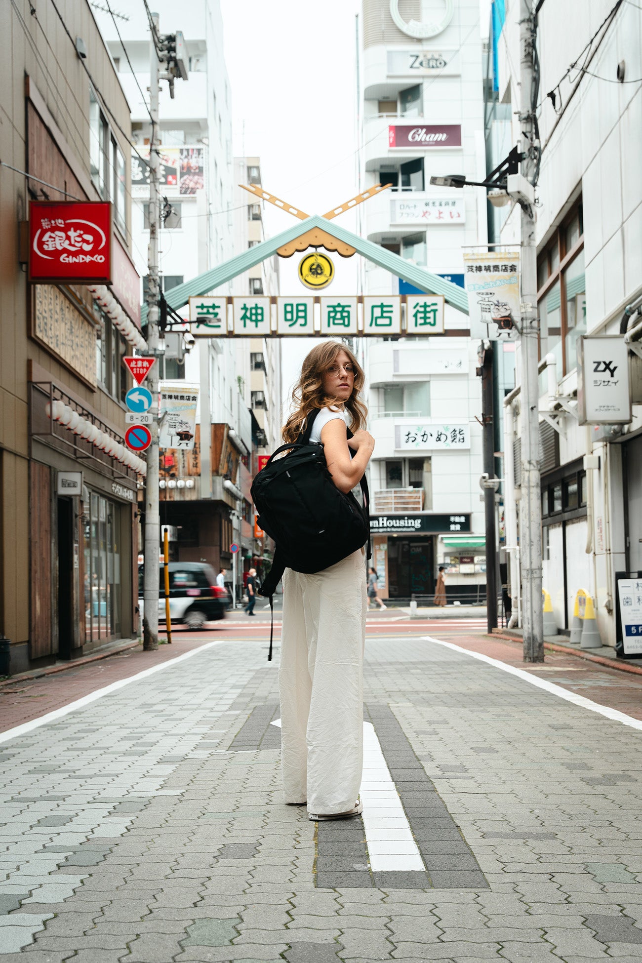 A woman stands on a city street wearing a white sleeveless top and wide-leg white pants, carrying a large black bag. The street has Japanese signage overhead and storefronts on both sides.